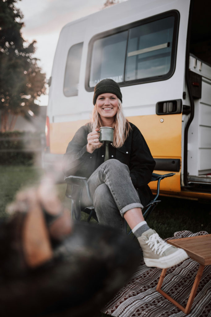 Femme assise devant un camping car dans la nature, tenant une tasse