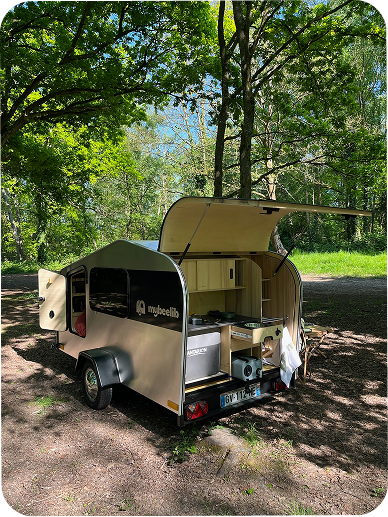 Tear Drop en forêt, hayon arrière relevé pour la cuisine extérieure, visuel “Nouvelle façon de voyager” sur fond de sous-bois ensoleillé.