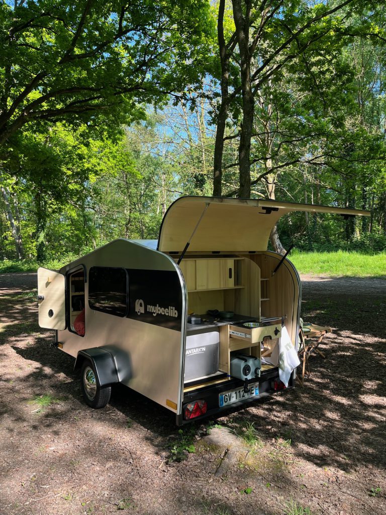 Tear Drop garée sous les arbres, cuisine arrière déployée avec plan de travail et rangements, scène de bivouac en pleine nature.