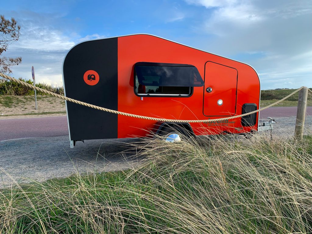 eardrop caravane BEEZARD rouge et noire près des dunes, vue latérale avec fenêtre, attelage et herbes hautes au premier plan.