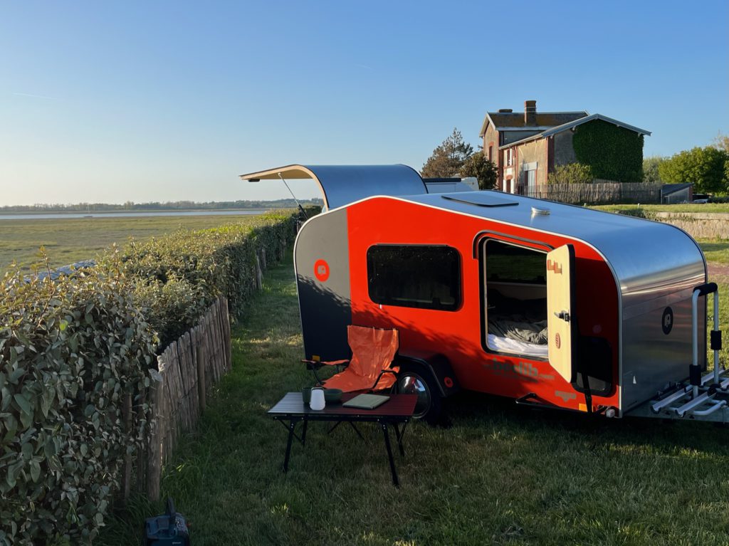 Tear Drop cuisine arrière au bord de l’eau : tiroir ouvert et plan de travail face au coucher de soleil, fauteuil de camping orange dans l’herbe.