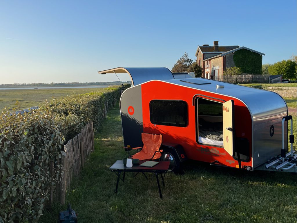 Tear Drop rouge et grise modèle BEEZARD, porte ouverte sur le couchage, installée en bord de mer avec étiquette “22 900 € tout équipé”.