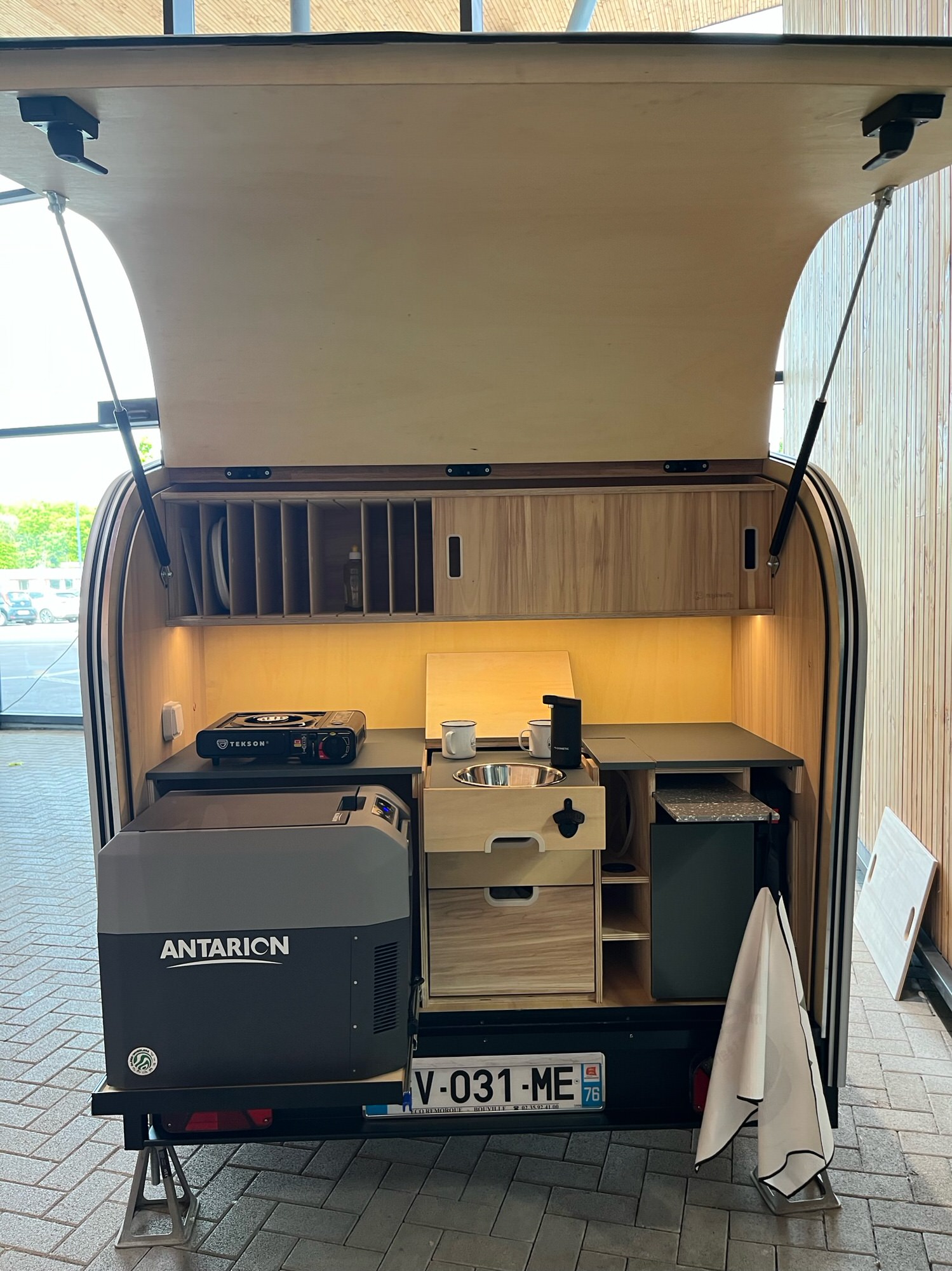 Cuisine arrière d’une mini-caravane : rangements en bois, plan de travail, réchaud, évier inox et frigo portable, éclairage chaud sous meuble.