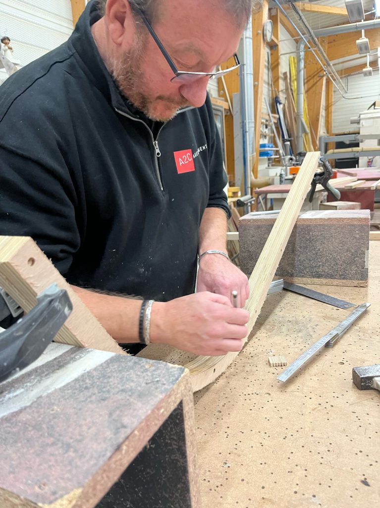 Artisan en atelier traçant et ajustant une pièce en bois pour une teardrop caravane, travail de précision sur établi avec outils de menuiserie.