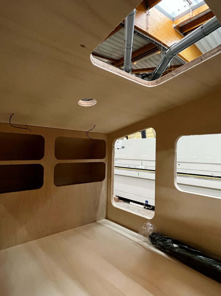 Intérieur brut d’une teardrop caravane en cours de fabrication : coque en bois avec niches de rangement, fenêtres découpées et ouverture de lanterneau au plafond.