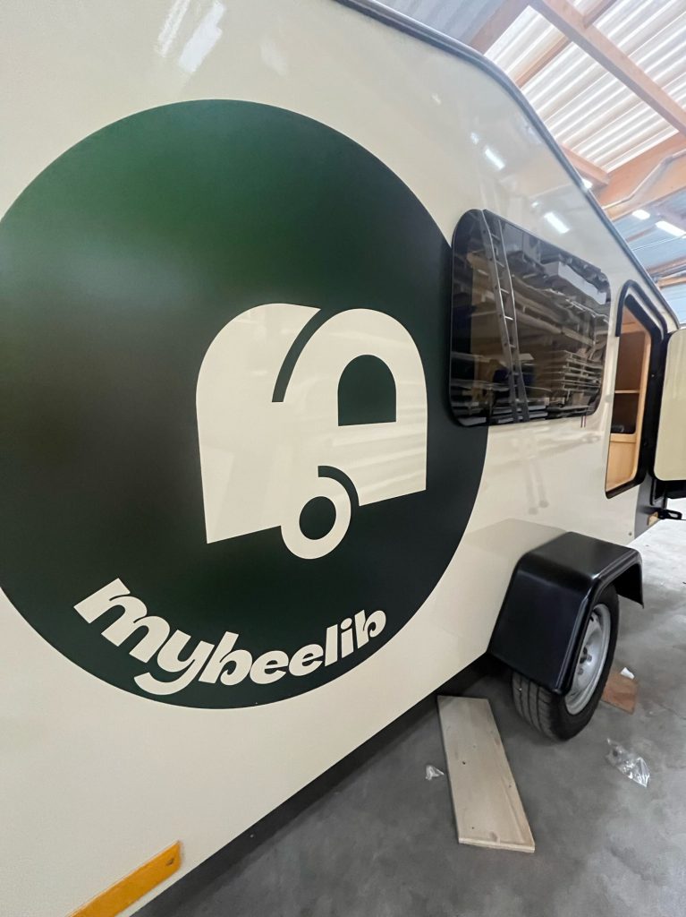 Tear Drop MyBeelib en atelier : gros plan sur le flanc avec logo rond et fenêtre teintée, carrosserie claire et roue apparente.