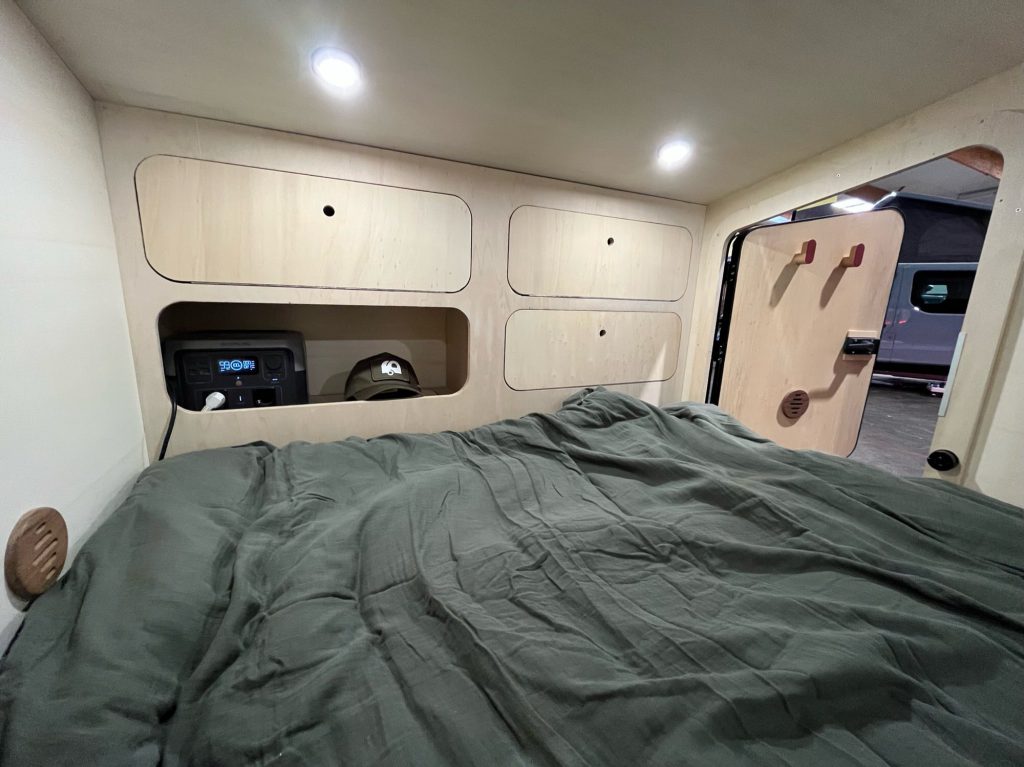 Couchage dans une teardrop caravane : lit double avec couette vert foncé, rangements intégrés au-dessus, éclairage LED et porte entrouverte.