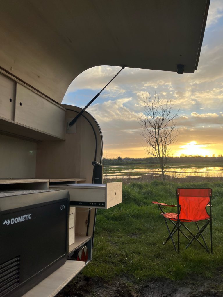 Cuisine arrière d’une teardrop caravane face au coucher de soleil : tiroir ouvert, plan de travail et glacière electrique, fauteuil de camping orange devant un plan d’eau.