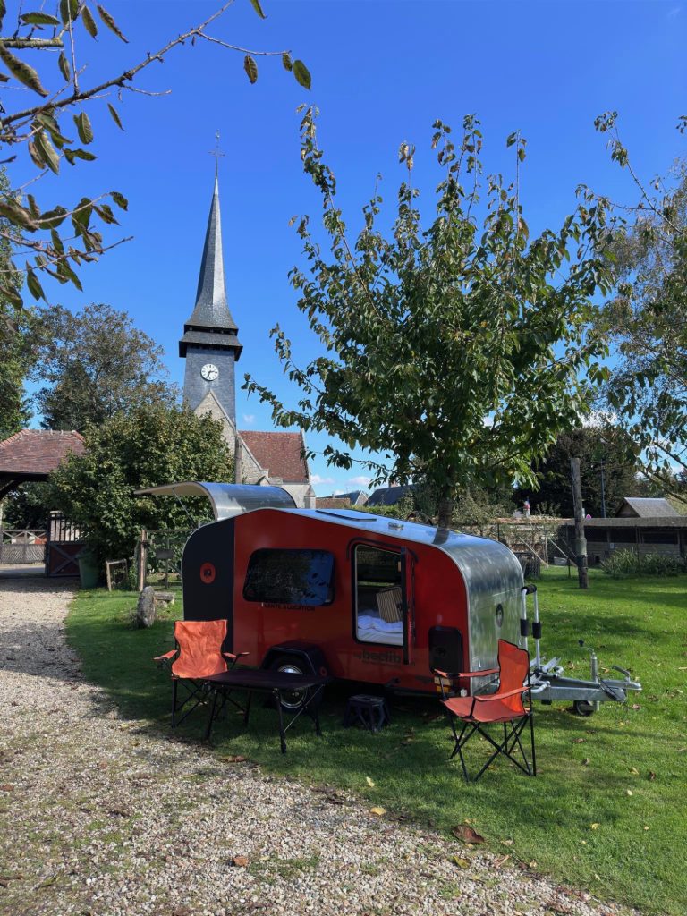 Tear Drop MyBeelib rouge et grise installée sur l’herbe près d’une église de village, coin camping avec table et chaises pliantes sous un ciel bleu.